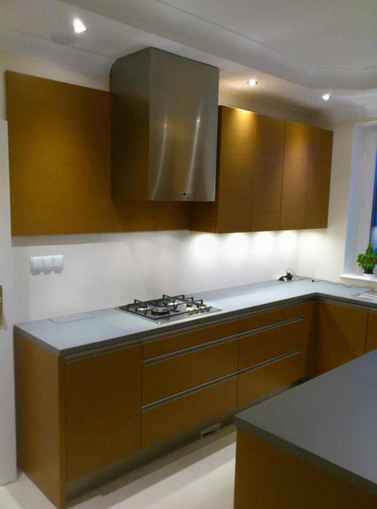 Nowoczesna kuchnia z drewnianymi szafkami i szarym blatem, oświetlona punktowymi lampami. Widoczny fragment okna i rośliny doniczkowej. Minimalistyczny design.