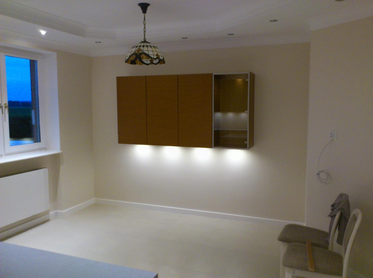 Nowoczesna aranżacja wnętrza z podświetlanymi szafkami wiszącymi w odcieniach brązu i beżu, oświetlonymi od dołu. Widoczne krzesła i lampa wisząca.