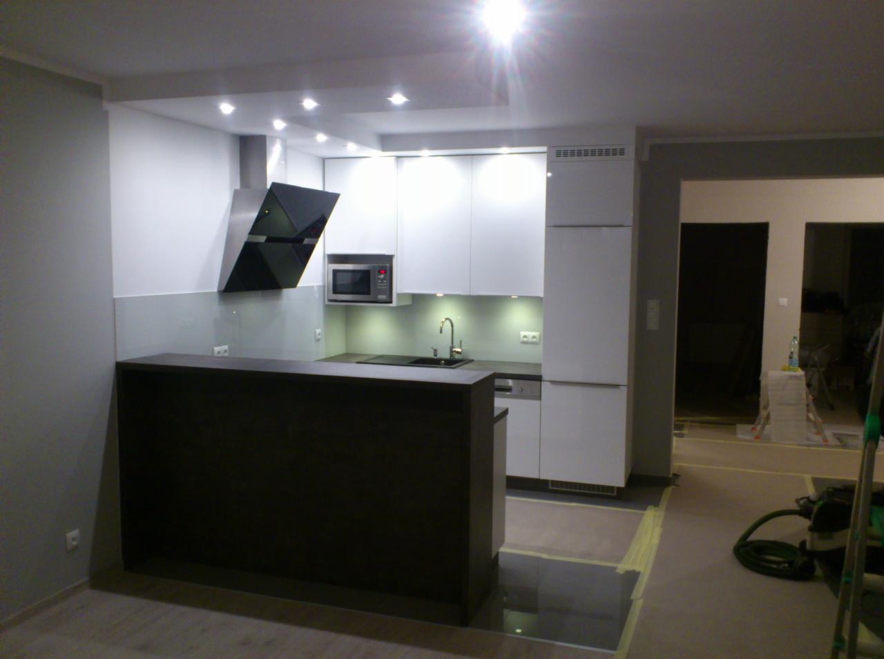 Nowoczesna, minimalistyczna kuchnia z białymi szafkami i czarnym okapem. Widoczny barek z ciemnego drewna, oświetlenie punktowe i sprzęt AGD.