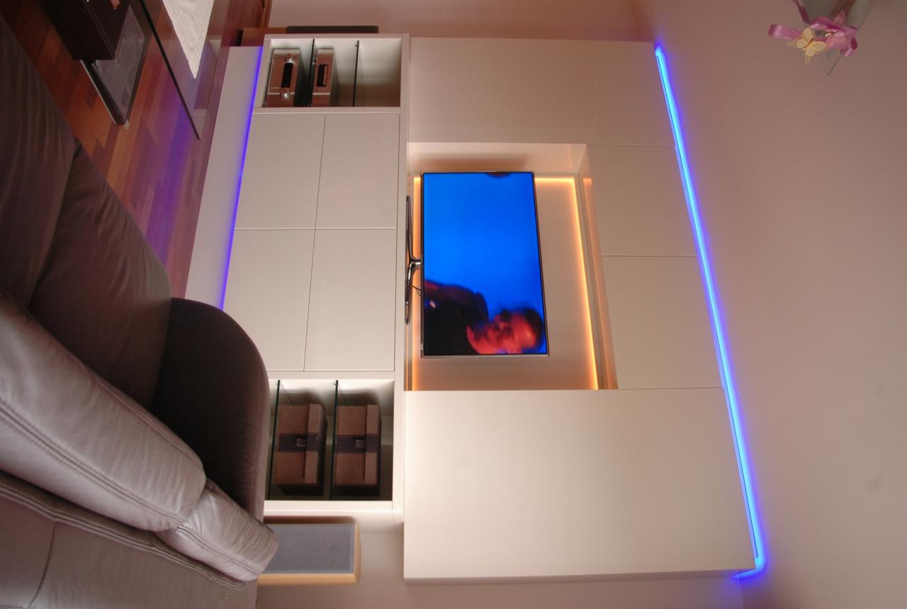Biała zabudowa meblowa na ścianie z wnęką na telewizor, podświetlona niebieskim LED i ciepłym światłem. W tle fragment sofy.