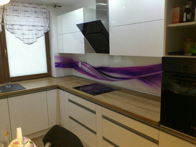 Nowoczesna kuchnia z białymi frontami, drewnianym blatem i fioletowym panelem dekoracyjnym. Widoczny okap i okno z firanką.