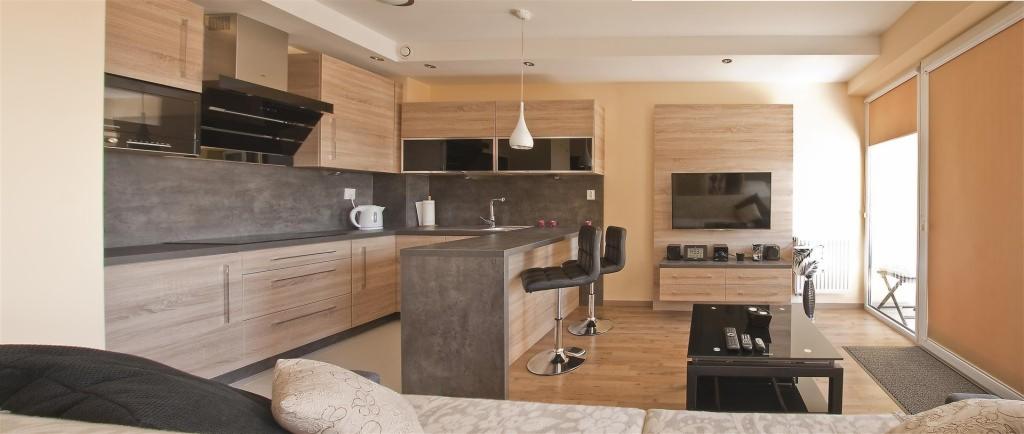 Jasna kuchnia z drewnianymi szafkami i szarym blatem, połączona z salonem. Widoczne hokery przy wyspie kuchennej i telewizor na ścianie.