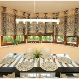 Jadalnia z oknem wykuszowym i roletami rzymskimi w geometryczny wz&oacute;r. St&oacute;ł z bieżnikiem w paski i plecionymi podkładkami. Nowoczesna lampa wisząca.