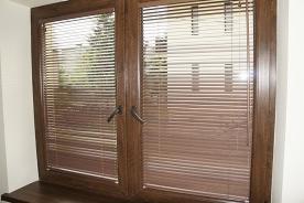 Brązowe okno z drewnianą ramą i zamontowanymi poziomymi żaluzjami w kolorze zbliżonym do ramy okiennej.