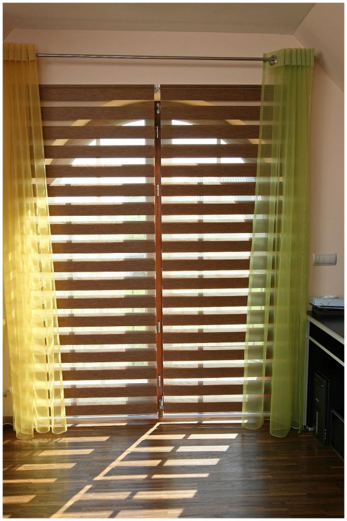 Okno z żaluzjami dzień-noc w odcieniach brązu, udekorowane żółtą i zieloną, zwiewną firanką, oświetlone promieniami słońca padającymi na parkiet.