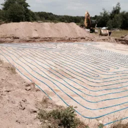 Fundament pod budynek z ułożonymi niebieskimi rurami ogrzewania podłogowego, w tle koparka i usypana ziemia.