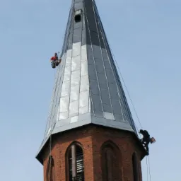 Dwóch alpinistów zabezpieczonych linami pracuje na wysokości, malując iglicę kościoła z cegły, pokrytą metalową blachą w słoneczny dzień.