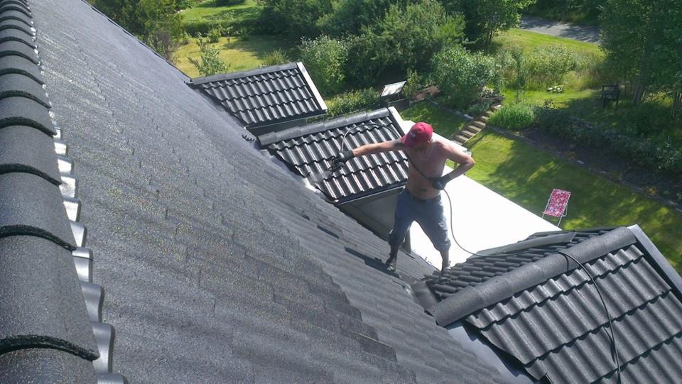 Mężczyzna w czerwonej czapce myje dachówkę ciśnieniowo. Część dachu jest już czysta, ukazując różnicę. Widoczny ogród z leżakiem w tle.