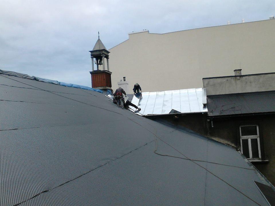 Prace na dachu z blachy trapezowej. Dwóch dekarzy w uprzężach montuje elementy na dachu budynku. W tle wieża kościelna i fragmenty innych budynków.