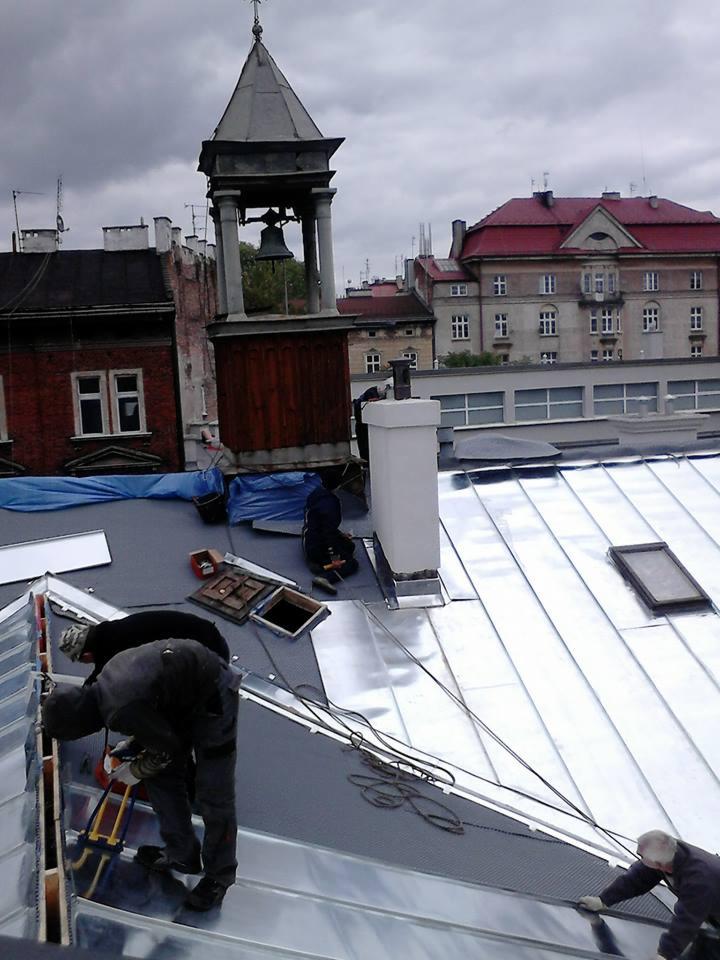 Montaż srebrnej blachy na dachu budynku. W tle wieża z dzwonem i zabudowa miejska pod pochmurnym niebem. Widoczne narzędzia i ekipa pracująca.