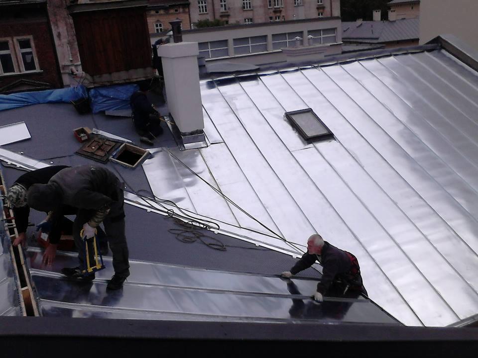 Montaż metalowej blachy na dachu budynku przez dekarzy. Widoczne narzędzia i elementy mocujące. Dach pokryty połyskującą blachą, z oknem dachowym.
