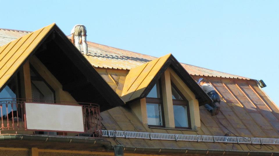 Prace na dachu domu z oknami mansardowymi. Dwóch pracowników na dachu, jeden w górnej części, drugi przy oknie. Dach pokryty blachą, widoczne elementy mocujące.