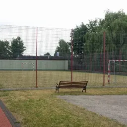 Ogrodzone, trawiaste boisko sportowe z bramkami, ławką i ścieżką z kostki, otoczone zielenią i budynkami. Czerwone słupki ogrodzenia.