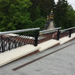Metalowa balustrada z elementami dekoracyjnymi, widok z g&oacute;ry na chodnik z kostki brukowej, w tle zieleń i fragment wieży z kopułą.
