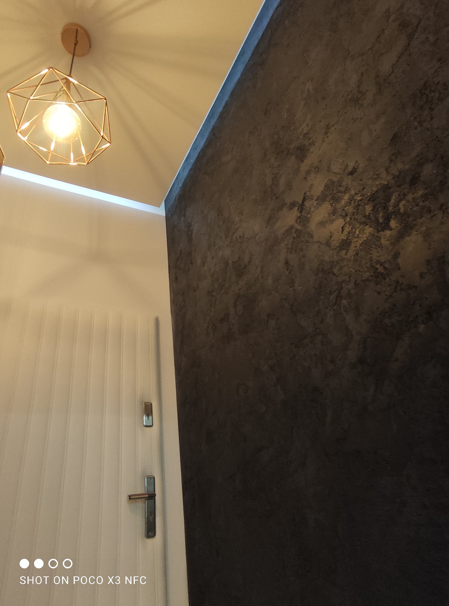 Dekoracyjna ściana z efektem betonu w ciemnych odcieniach szarości sąsiadująca z białą, panelową ścianą i złotą lampą sufitową.