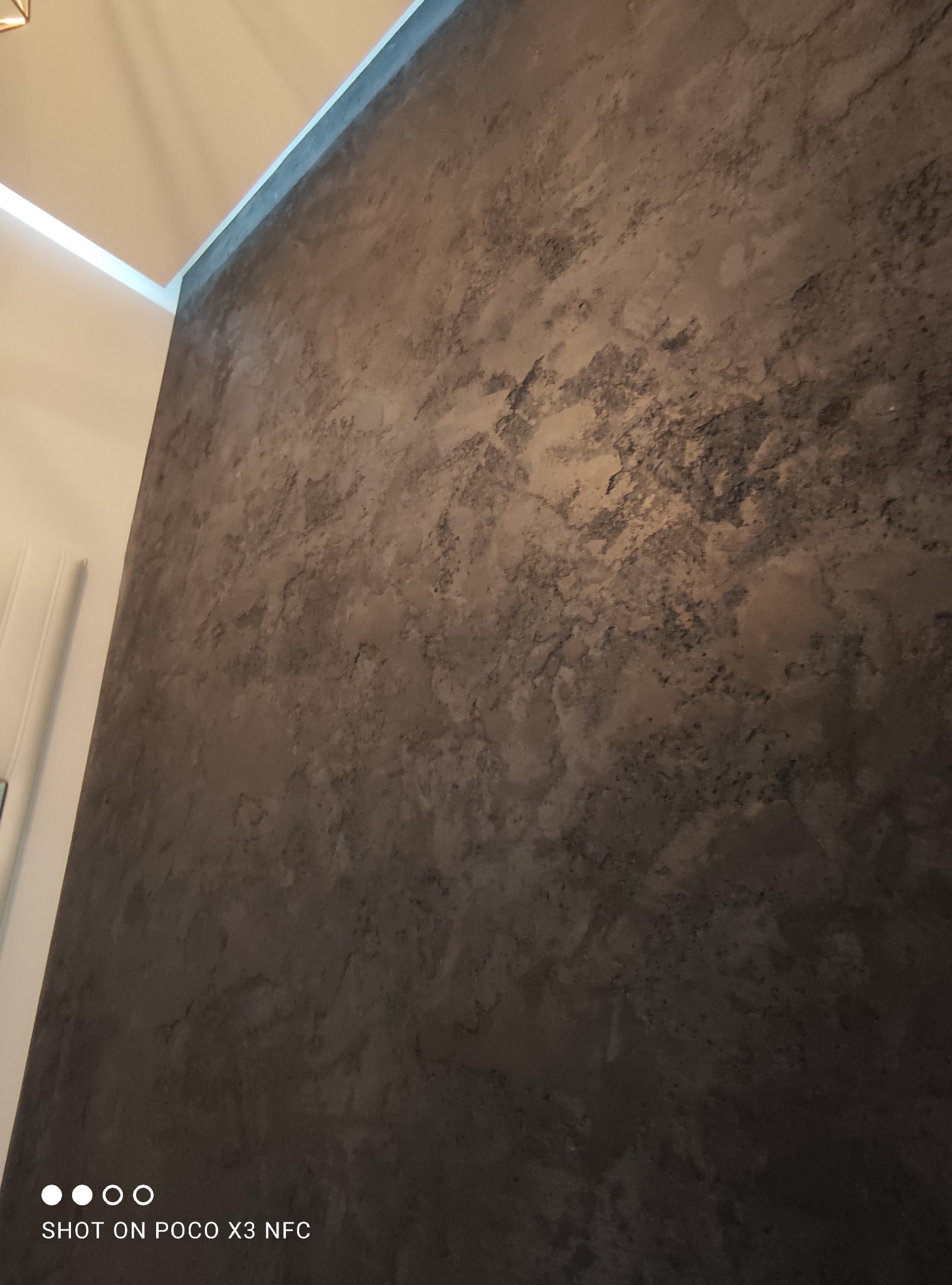 Ściana wewnętrzna z efektem dekoracyjnym betonu architektonicznego, oświetlona od góry, widoczny fragment sufitu i listwy przypodłogowej.