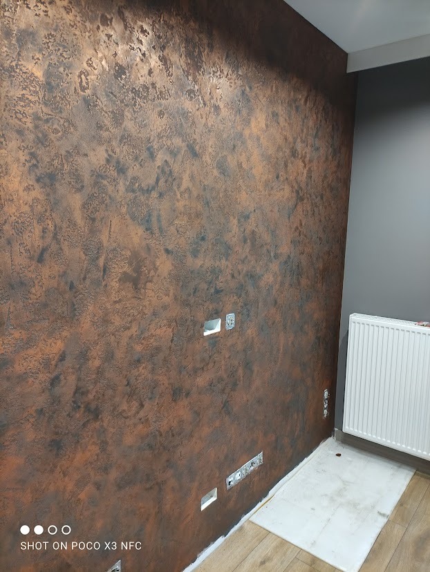 Ściana z dekoracyjną fakturą w odcieniach brązu i czerni, widoczne puszki instalacyjne bez osprzętu, grzejnik i fragment podłogi.
