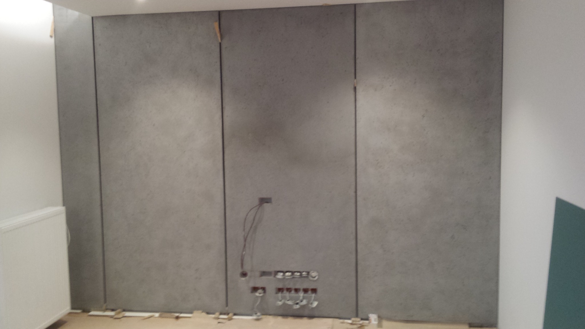 Ściana wykończona panelami imitującymi beton, z widocznymi puszkami elektrycznymi i przewodami, w trakcie prac wykończeniowych.