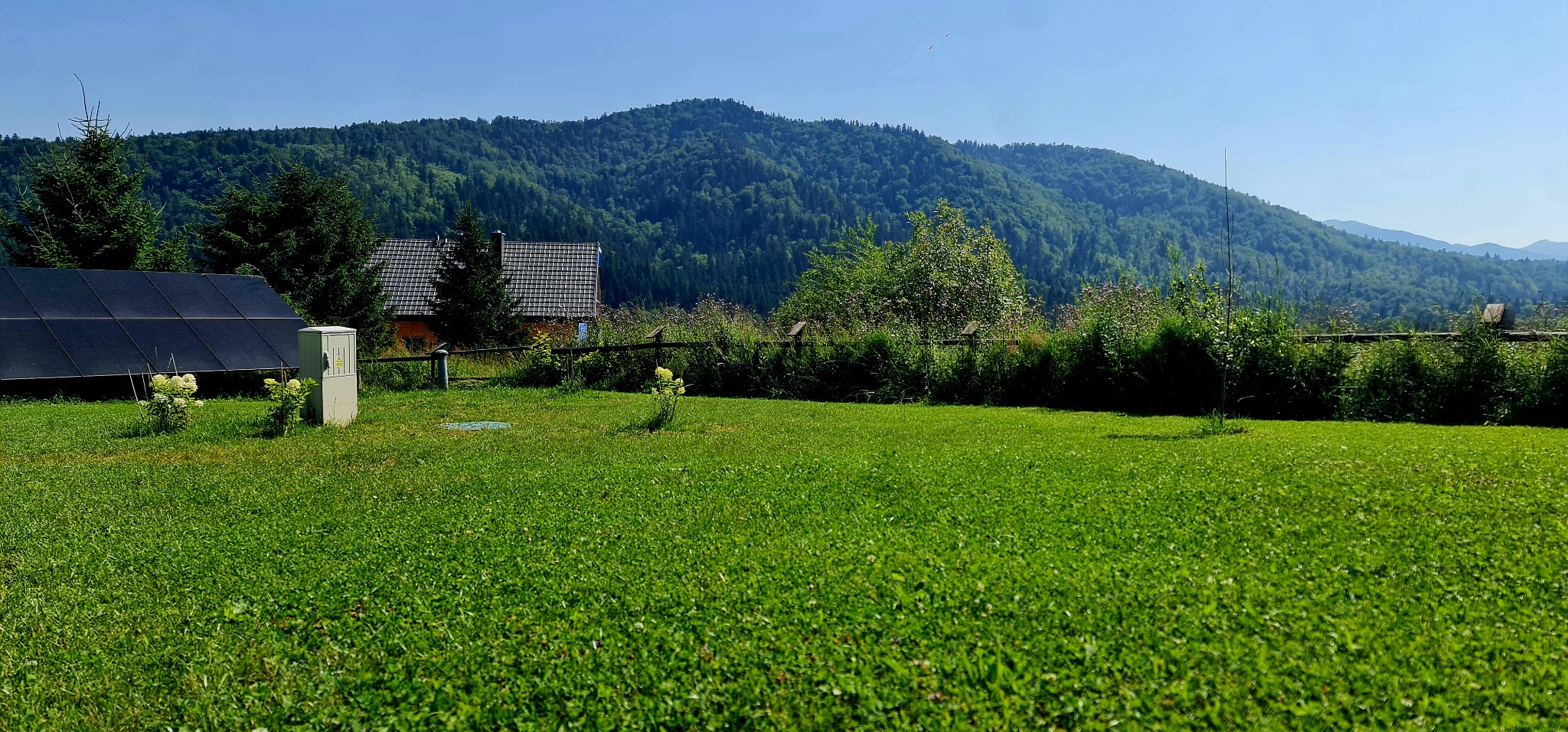 Panel słoneczny i skrzynka elektryczna na zielonym trawniku z widokiem na zalesione wzgórza w tle. Ujęcie w słoneczny dzień.