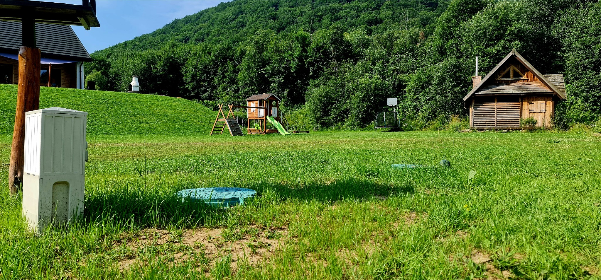 Zielony teren z pokrywą studzienki i skrzynką elektryczną w Kunowie. W tle domek, plac zabaw i zalesione wzgórze. Słoneczny dzień, trawa, rekreacja.