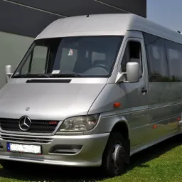 Srebrny minibus Mercedes-Benz Sprinter z przyciemnianymi szybami, zaparkowany na trawniku przed budynkiem o szarej elewacji.