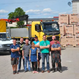 Grupa pracowników budowlanych pozuje na tle dwóch samochodów dostawczych i palet z cegłami marki Roben na placu budowy.