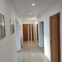 Korytarz z panelami podłogowymi w odcieniach beżu, białymi ścianami, drewnianymi drzwiami i oświetleniem punktowym w suficie. Na ścianach wiszą obrazy w srebrnych ramach.