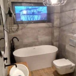 Nowoczesna łazienka w szarościach z drewnianymi akcentami, owalna wanna wolnostojąca, umywalka nablatowa na drewnianym blacie, wisząca toaleta, geometryczne lampy wiszące i okno z widokiem na drzewa.