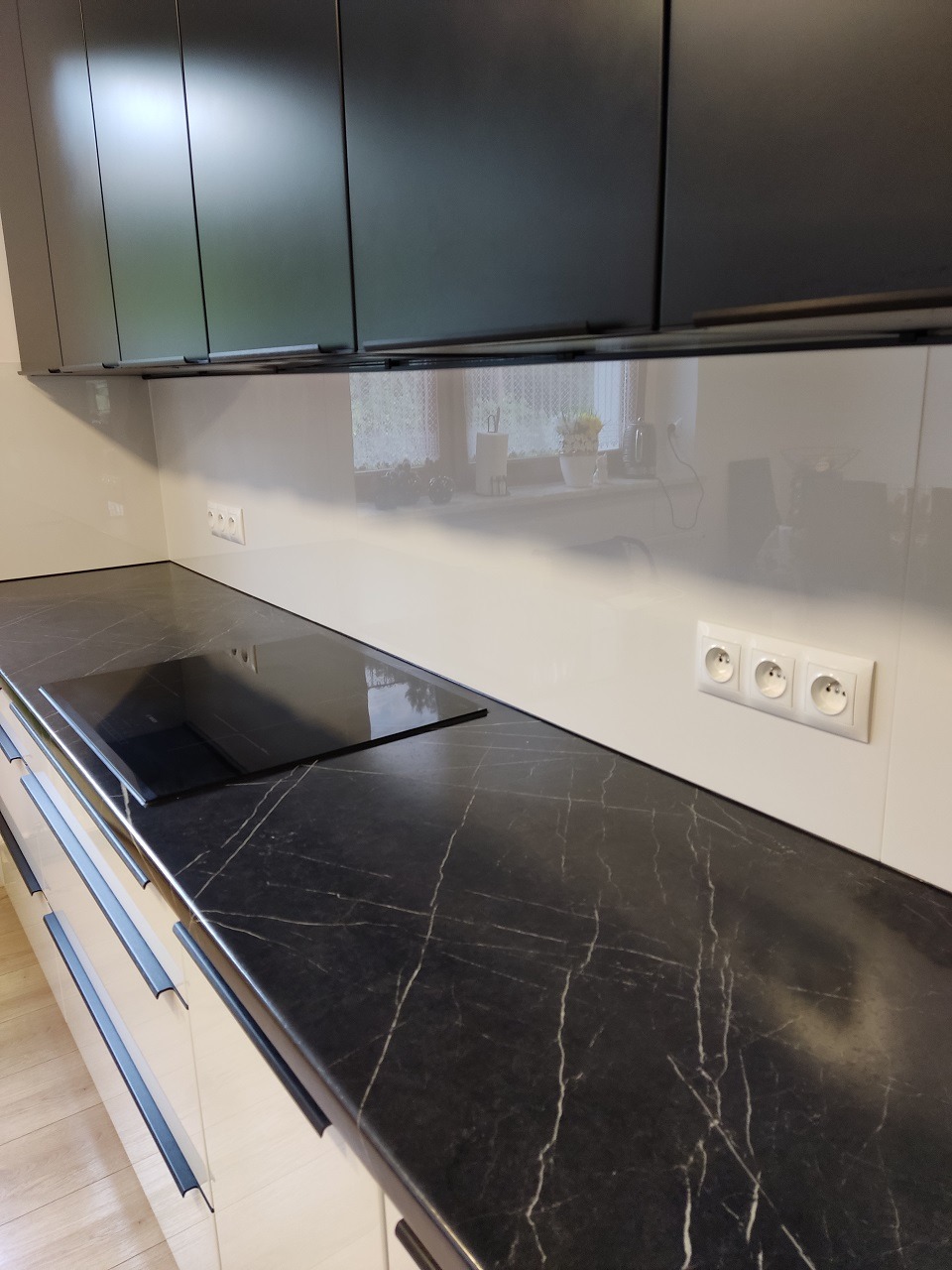 Nowoczesna kuchnia z czarnym blatem imitującym marmur, jasnymi szafkami z długimi, czarnymi uchwytami i szarymi szafkami wiszącymi. Widoczna płyta indukcyjna i gładka, jasna ściana nad blatem...