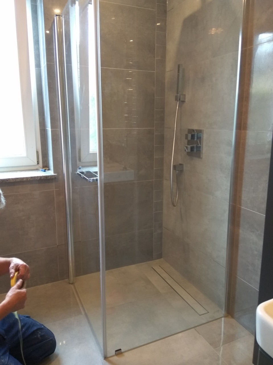 Szklana kabina prysznicowa z odpływem liniowym w trakcie montażu; widoczne płytki w odcieniach szarości i częściowo widoczna osoba z miarką.