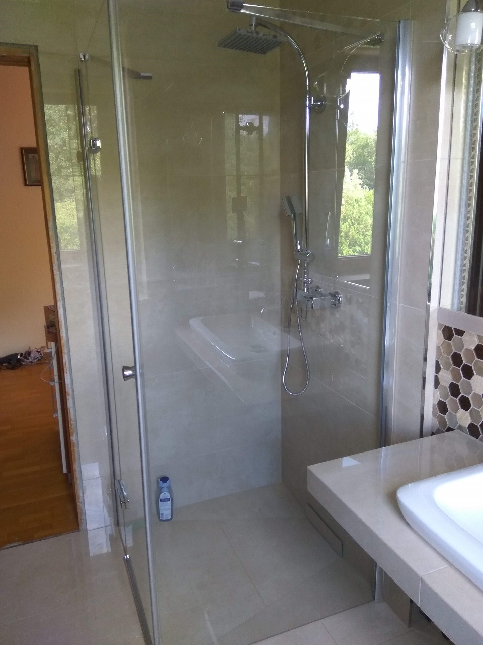 Nowoczesna kabina prysznicowa z deszczownicą i szklanymi drzwiami, widoczna butelka płynu do mycia pod prysznicem, beżowe płytki i heksagonalna mozaika na ścianie obok umywalki.