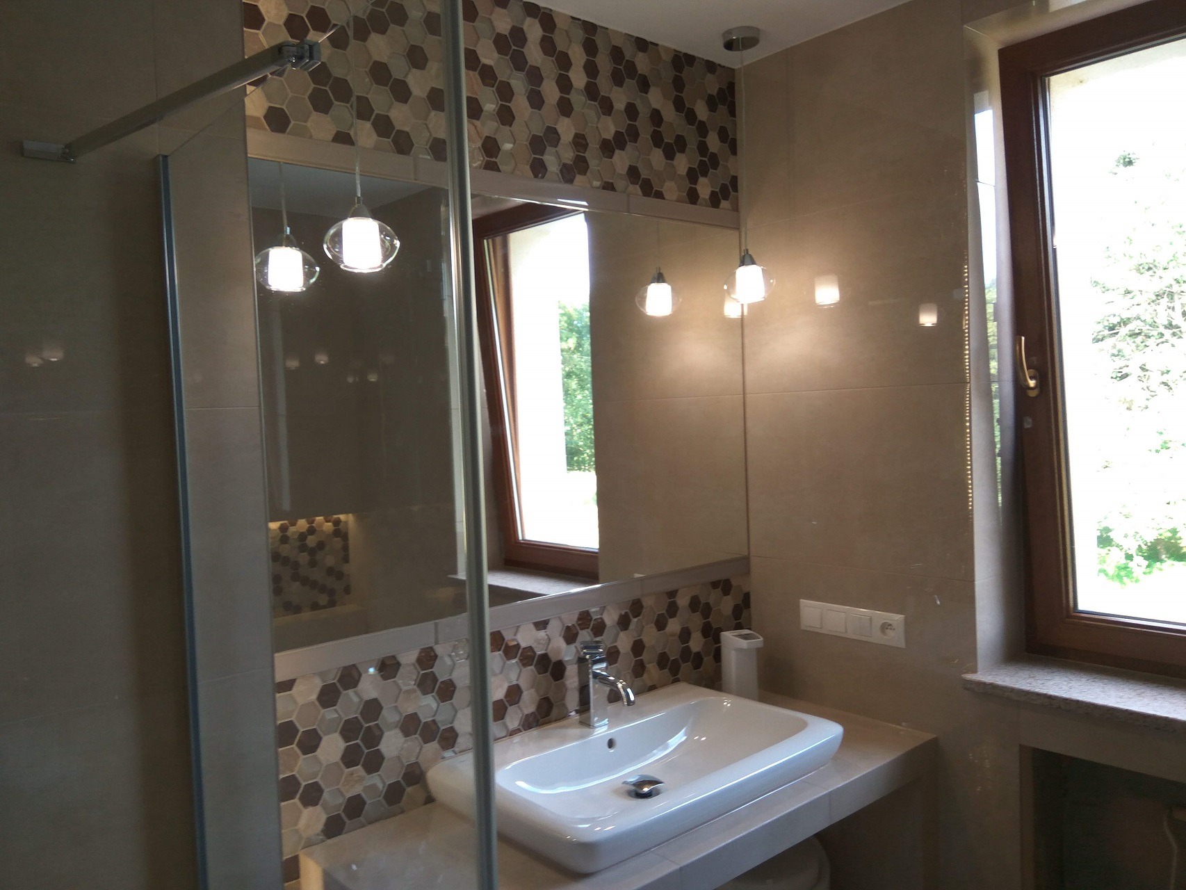 Ukończona łazienka z kabiną prysznicową, umywalką nablatową i mozaiką heksagonalną w odcieniach brązu i beżu, oświetlona wiszącymi lampami kulami.