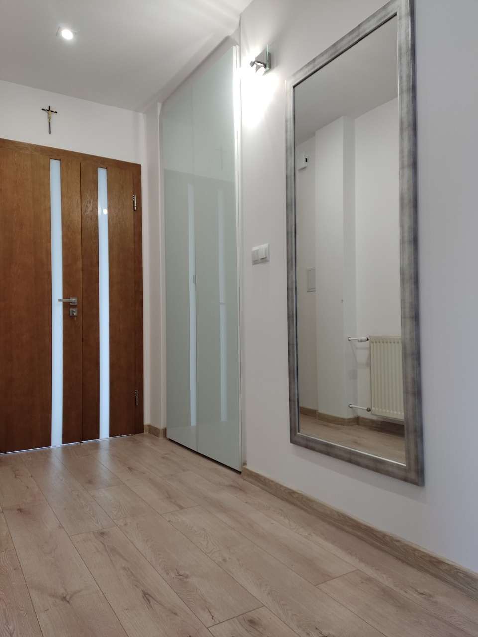 Jasne wnętrze przedpokoju z drewnianą podłogą, lustrem w srebrnej ramie, przeszkloną szafą w zabudowie i drewnianymi drzwiami z białymi wstawkami.