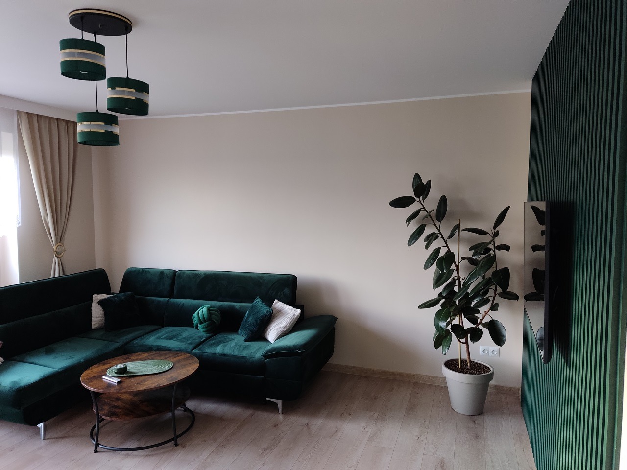 Nowoczesny salon z zieloną sofą narożną, okrągłym stolikiem kawowym, wiszącą lampą z trzema zielonymi kloszami i ścianą z zielonych lameli, uzupełniony o roślinę doniczkową.