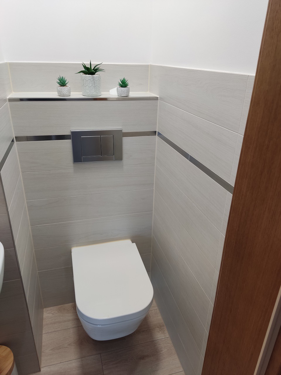 Nowoczesna toaleta podwieszana w wykończonej łazience, z płytkami imitującymi drewno, srebrnymi akcentami i trzema małymi roślinami doniczkowymi na półce nad spłuczką.