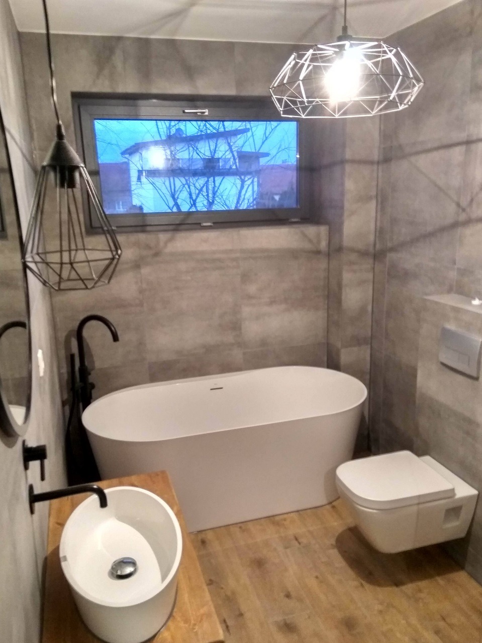 Nowoczesna łazienka w szarościach z drewnianymi akcentami, owalna wanna wolnostojąca, umywalka nablatowa na drewnianym blacie, wisząca toaleta, geometryczne lampy wiszące i okno z widokiem na drzewa.