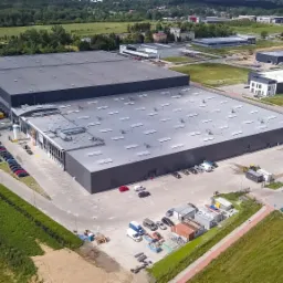 Zdjęcie z lotu ptaka przedstawia dużą, szarą halę magazynową z licznymi świetlikami dachowymi, zaparkowanymi samochodami osobowymi i ciężarowymi w strefie załadunku oraz otaczającą infrastrukturą...