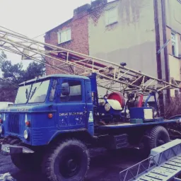 Niebieska ciężarówka wiertnicza z napisem 'Zakład Studniarski' na tle budynku mieszkalnego, z widoczną konstrukcją wiertniczą i elementami wyposażenia.