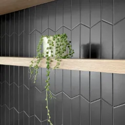 Ciemnoszare, błyszczące płytki ułożone w jodełkę na ścianie, z jasnymi drewnianymi półkami i zwisającą rośliną w białej doniczce.