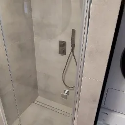 Szklana kabina prysznicowa z szarymi płytkami, zamontowana bateria podtynkowa z deszczownicą i słuchawką, obok pralka Toshiba.