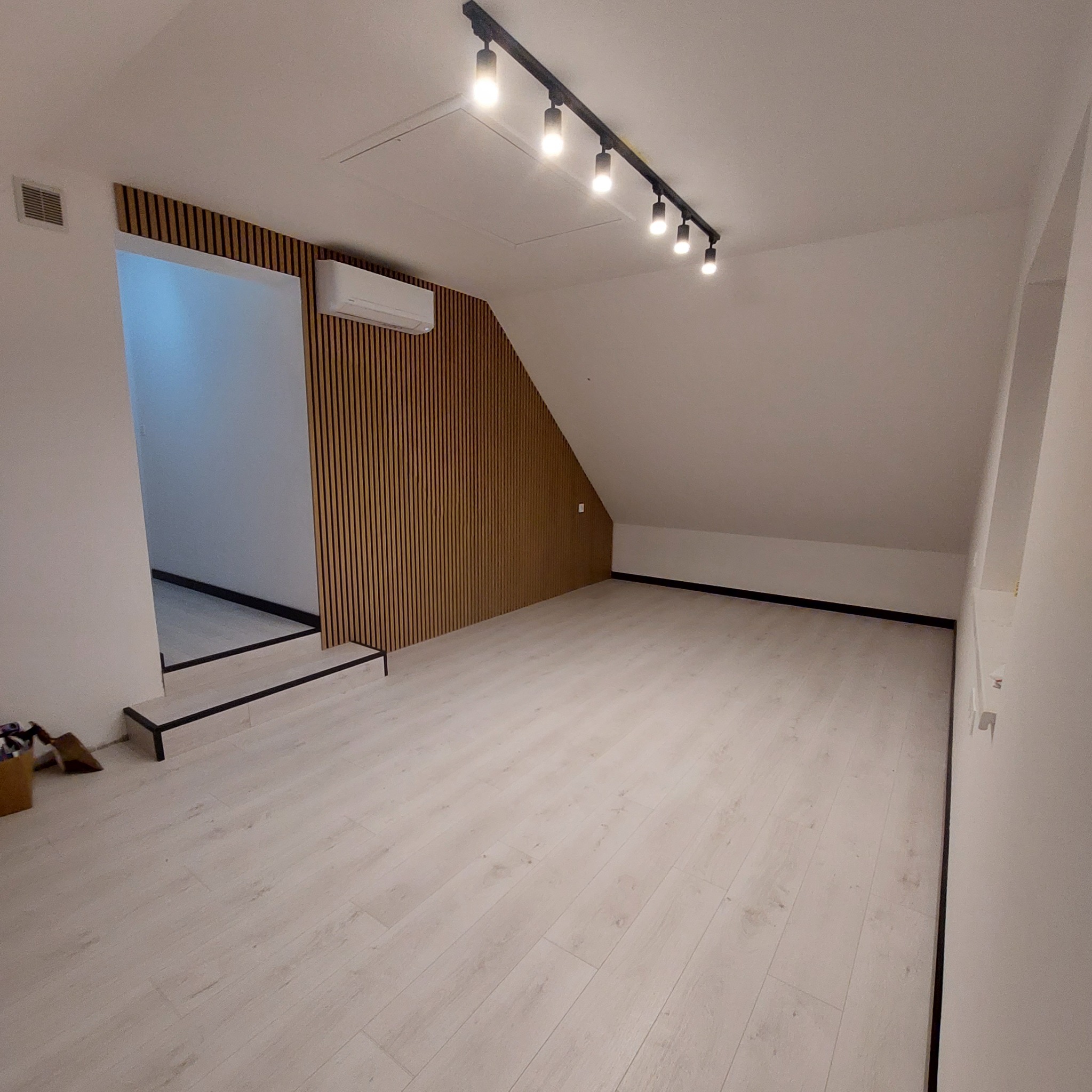 Poddasze po remoncie z drewnianą ścianą, klimatyzacją, oświetleniem szynowym i podłogą z paneli w jasnym odcieniu drewna.