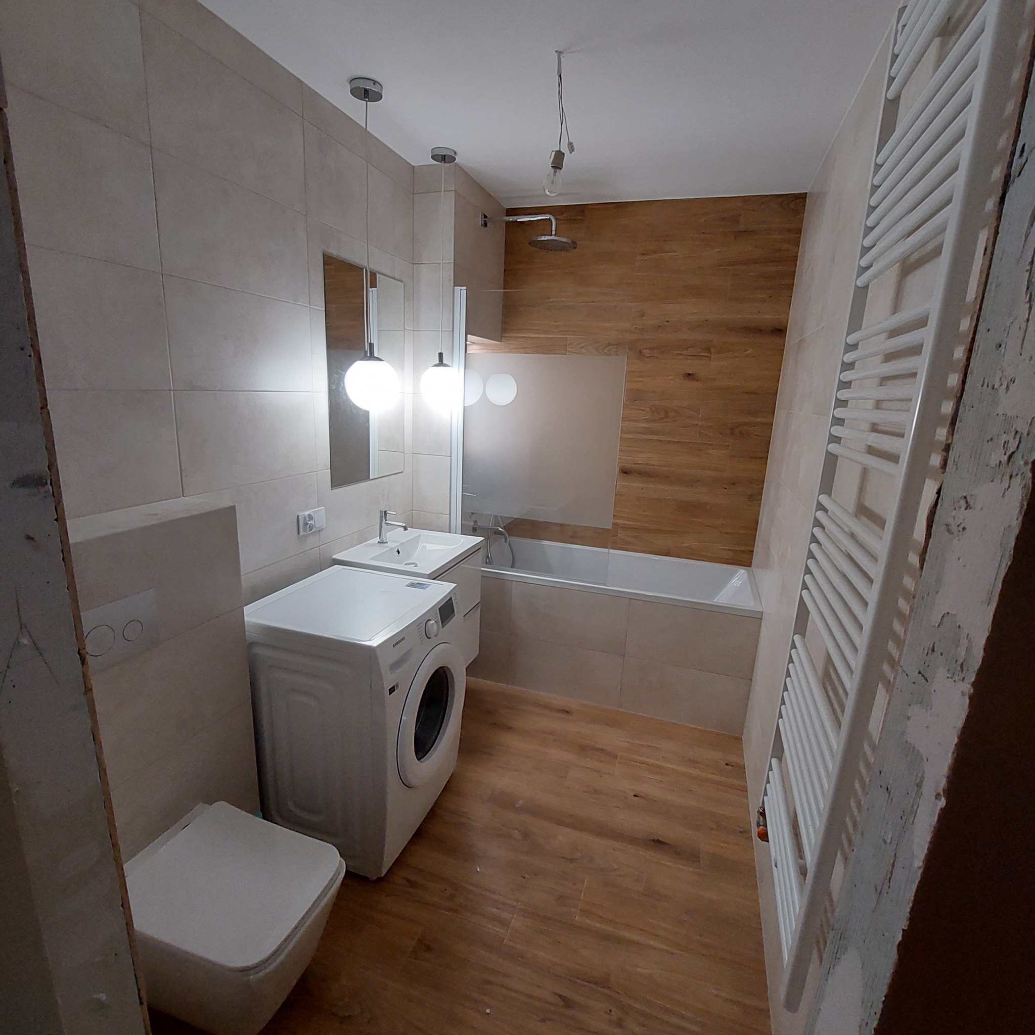 Nowoczesna łazienka po remoncie z drewnopodobnymi płytkami, białą pralką, umywalką z szafką, grzejnikiem drabinkowym i wiszącymi lampami kulami; widoczny fragment niedokończonej ściany.