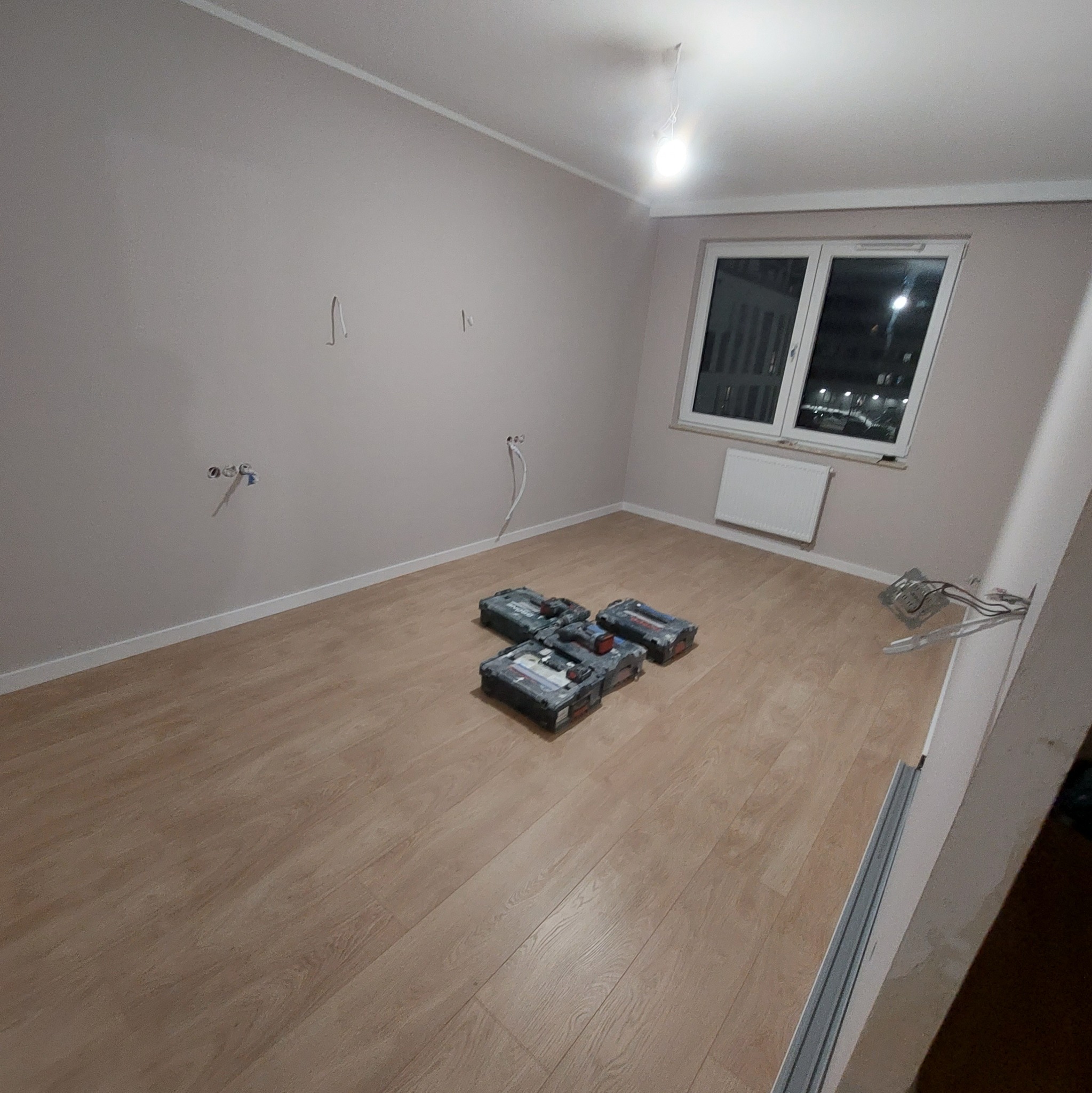 Pokój w trakcie prac wykończeniowych: świeżo położona podłoga, ściany w jasnym odcieniu z wystającymi przewodami elektrycznymi, skrzynki z narzędziami na podłodze, widok na okno.