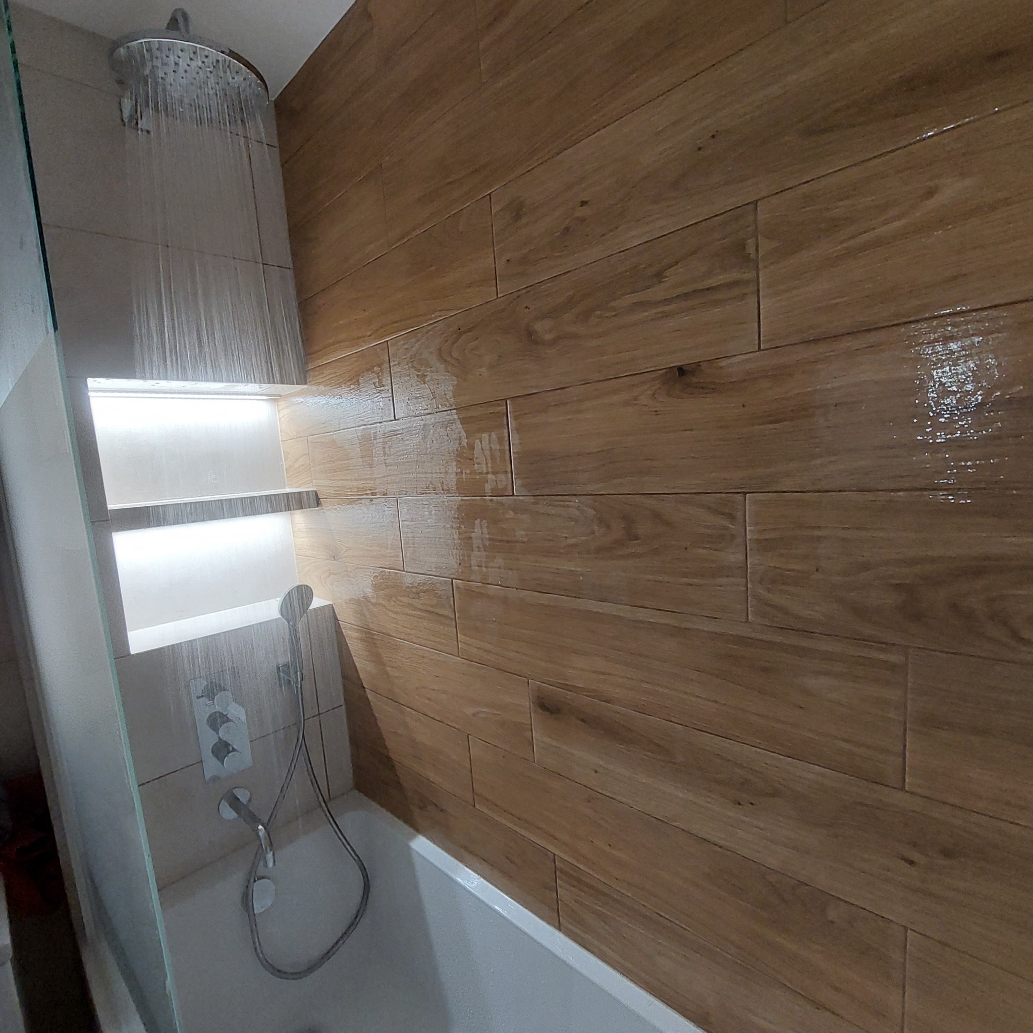 Nowoczesna łazienka z deszczownicą w trakcie użytkowania, drewnopodobne płytki na ścianie i podświetlana półka w kabinie prysznicowej.