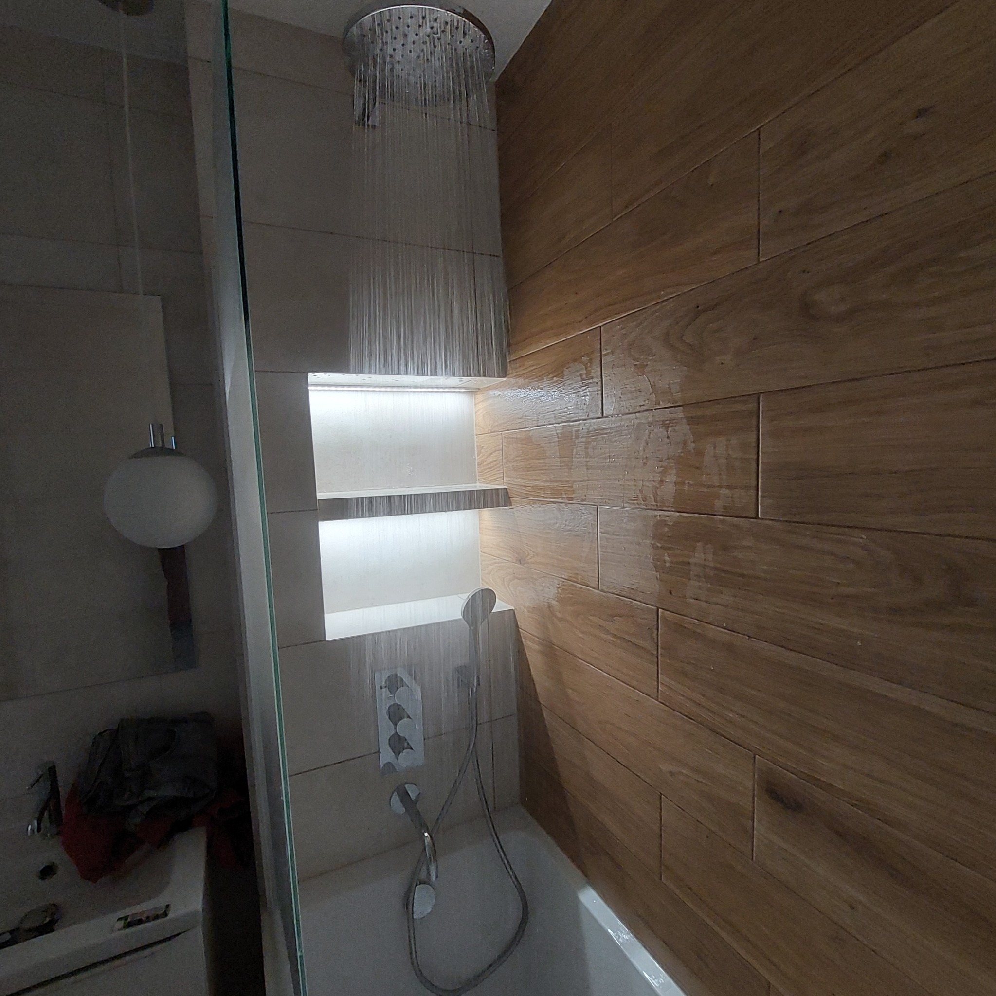 Nowoczesna łazienka z deszczownicą w trakcie działania, podświetlane półki w ścianie, drewnopodobne płytki na ścianie, bateria termostatyczna.