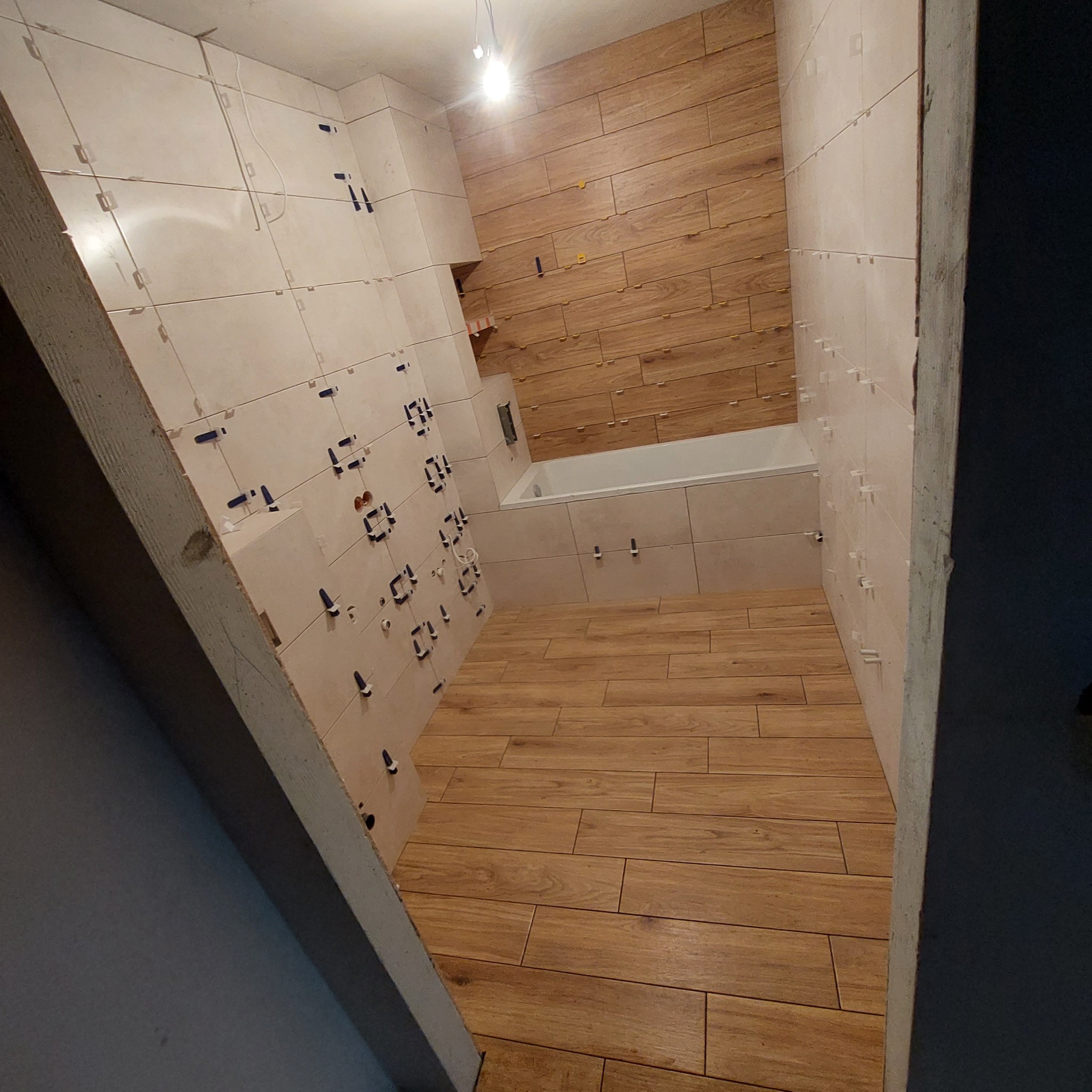 Łazienka w trakcie układania płytek podłogowych i ściennych, z widocznymi krzyżykami dystansowymi i poziomą okładziną przypominającą drewno na jednej ze ścian oraz białą wanną.