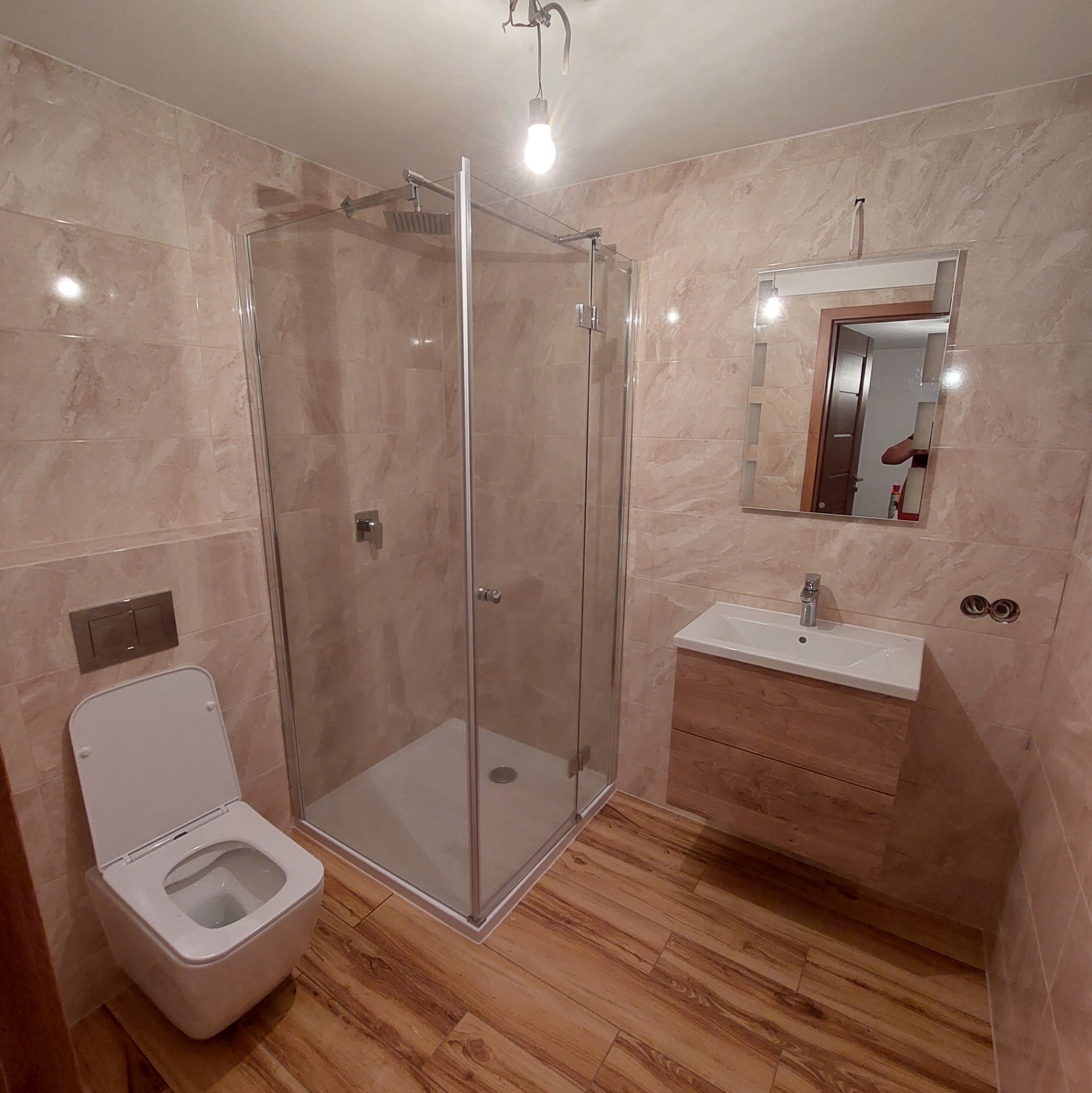 Wykończona łazienka z beżowymi płytkami, kabiną prysznicową ze szklanymi ścianami, podwieszaną toaletą, umywalką z szafką z drewnopodobnym fornirem i lustrem.