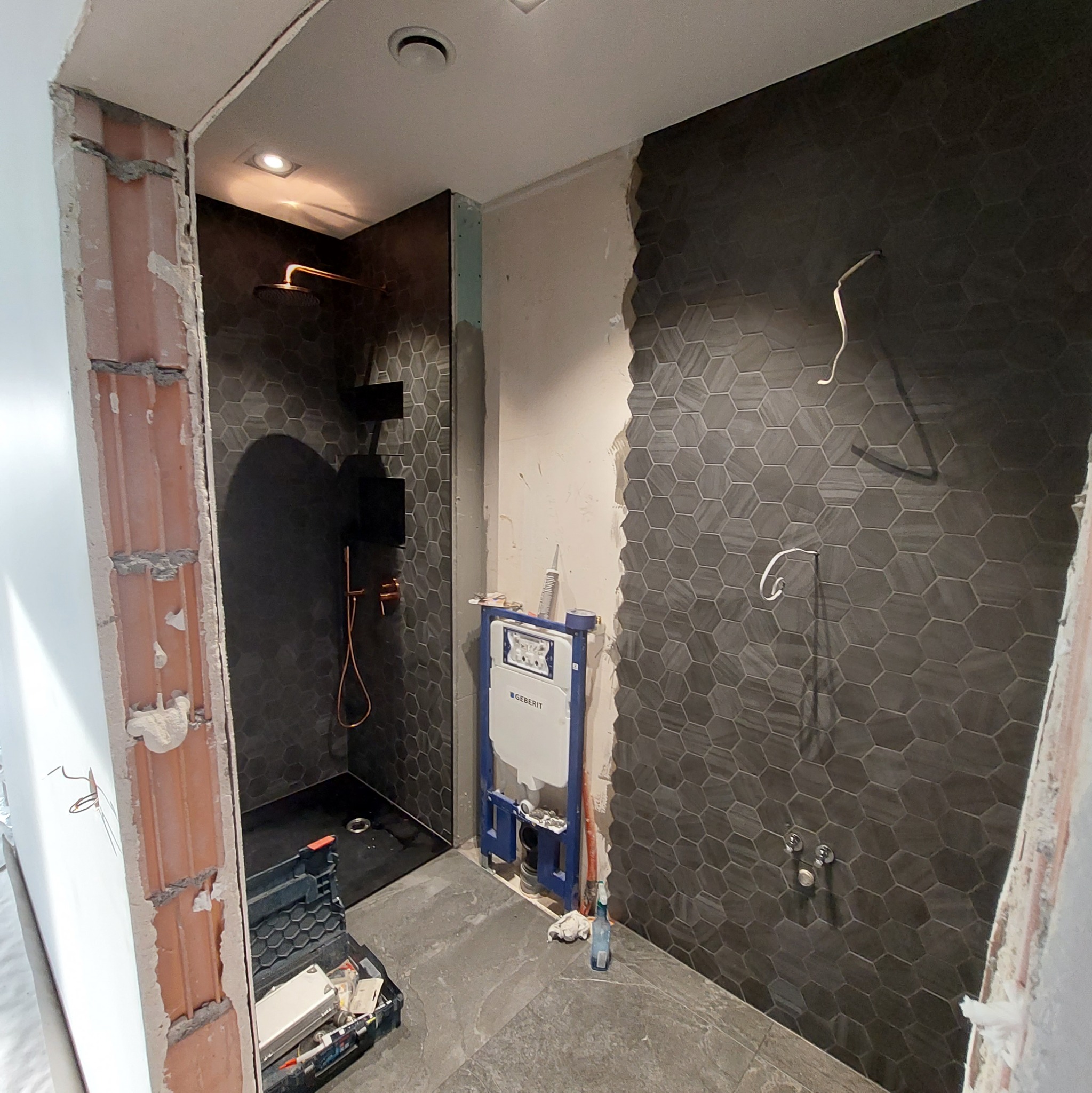 Łazienka w trakcie prac: heksagonalne płytki w odcieniach szarości, instalacja podtynkowa Geberit, widoczne przewody elektryczne i narzędzia.