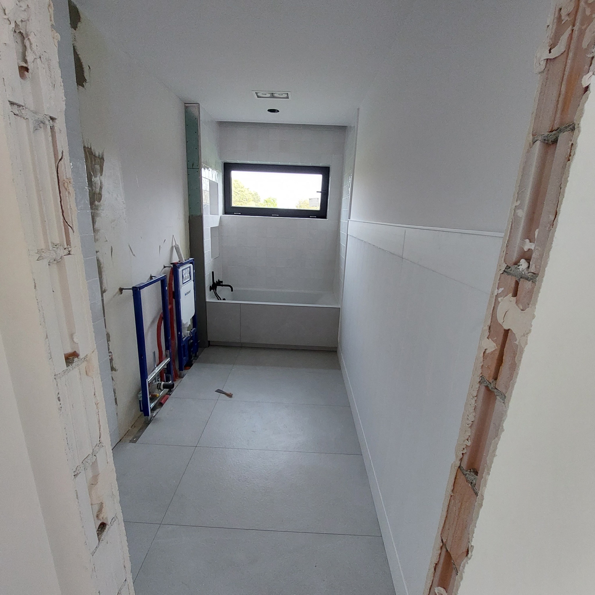 Wnętrze łazienki w trakcie prac: widoczne niedokończone ściany, zamontowana instalacja podtynkowa WC, położone płytki podłogowe i częściowo ścienne, okno nad wanną.