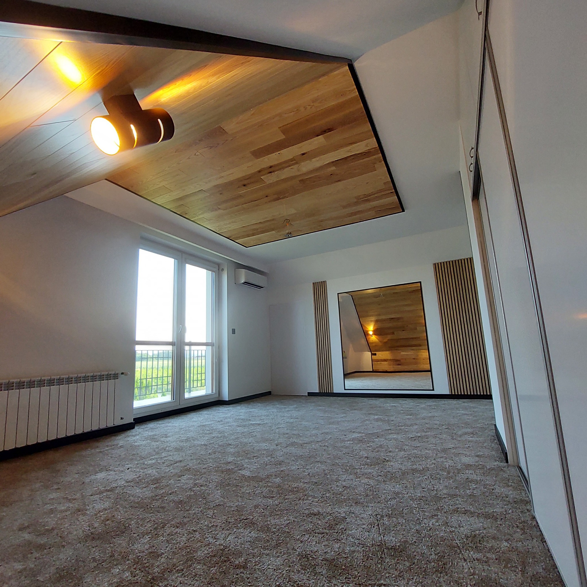 Przestronne, jasne wnętrze pokoju z drewnianym sufitem podwieszanym, oświetleniem punktowym, dużym oknem z widokiem na zieleń i lustrem powiększającym przestrzeń.